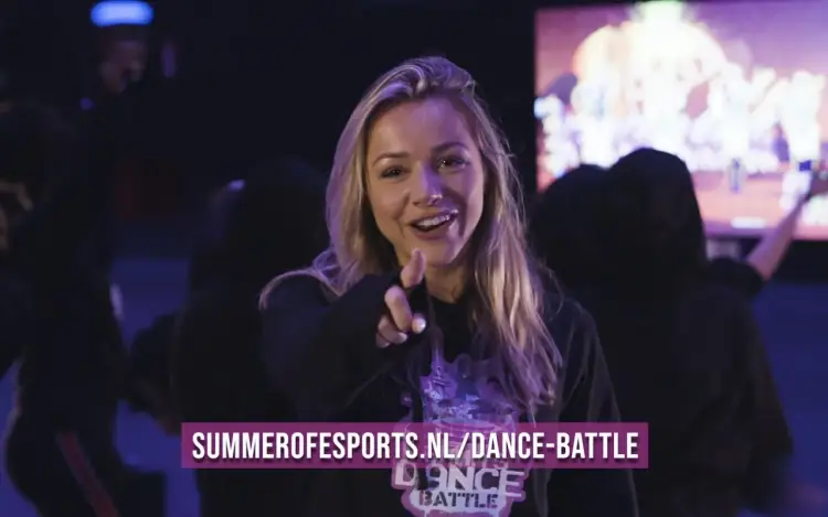 Een jonge vrouw glimlacht en wijst naar de camera tijdens een esports- of dansbattle-evenement.