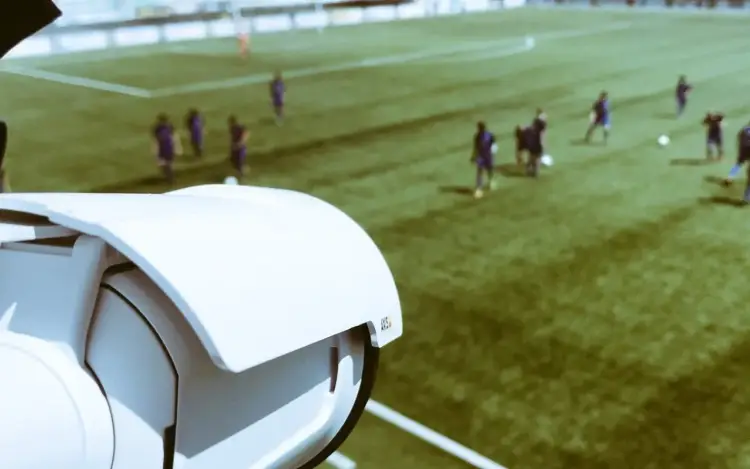 Close-up van een slimme sportcamera naast een voetbalveld waar spelers actief zijn.