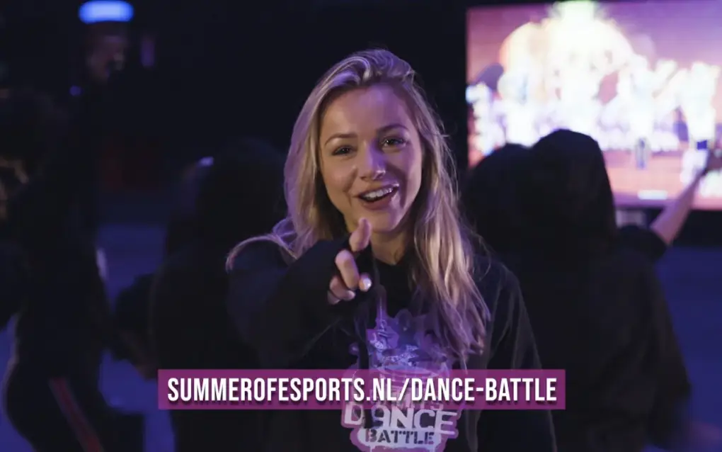 Een jonge vrouw glimlacht en wijst naar de camera tijdens een esports- of dansbattle-evenement.