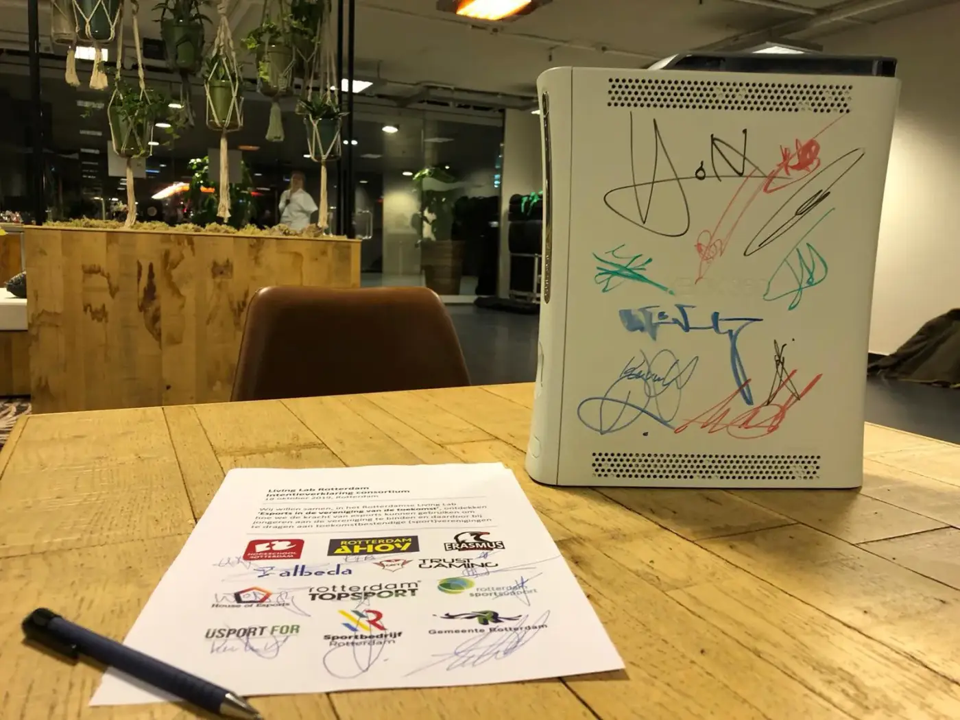 Een houten tafel met een ondertekend document van verschillende organisaties, naast een witte spelcomputer vol handtekeningen en tekeningen.