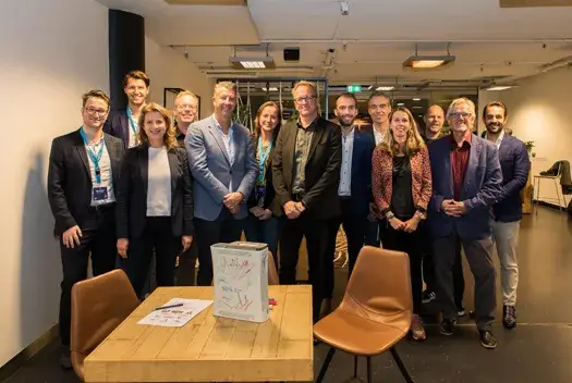 Groepsfoto van een tiental mensen in een vergaderruimte, lachend naar de camera, rondom een tafel.