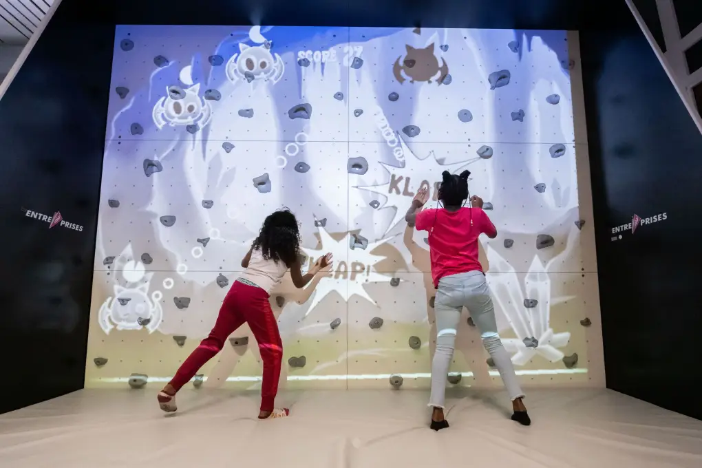 Een jongen en een meisje beklimmen een interactieve klimmuur waarop geanimeerde spel-elementen worden geprojecteerd.
