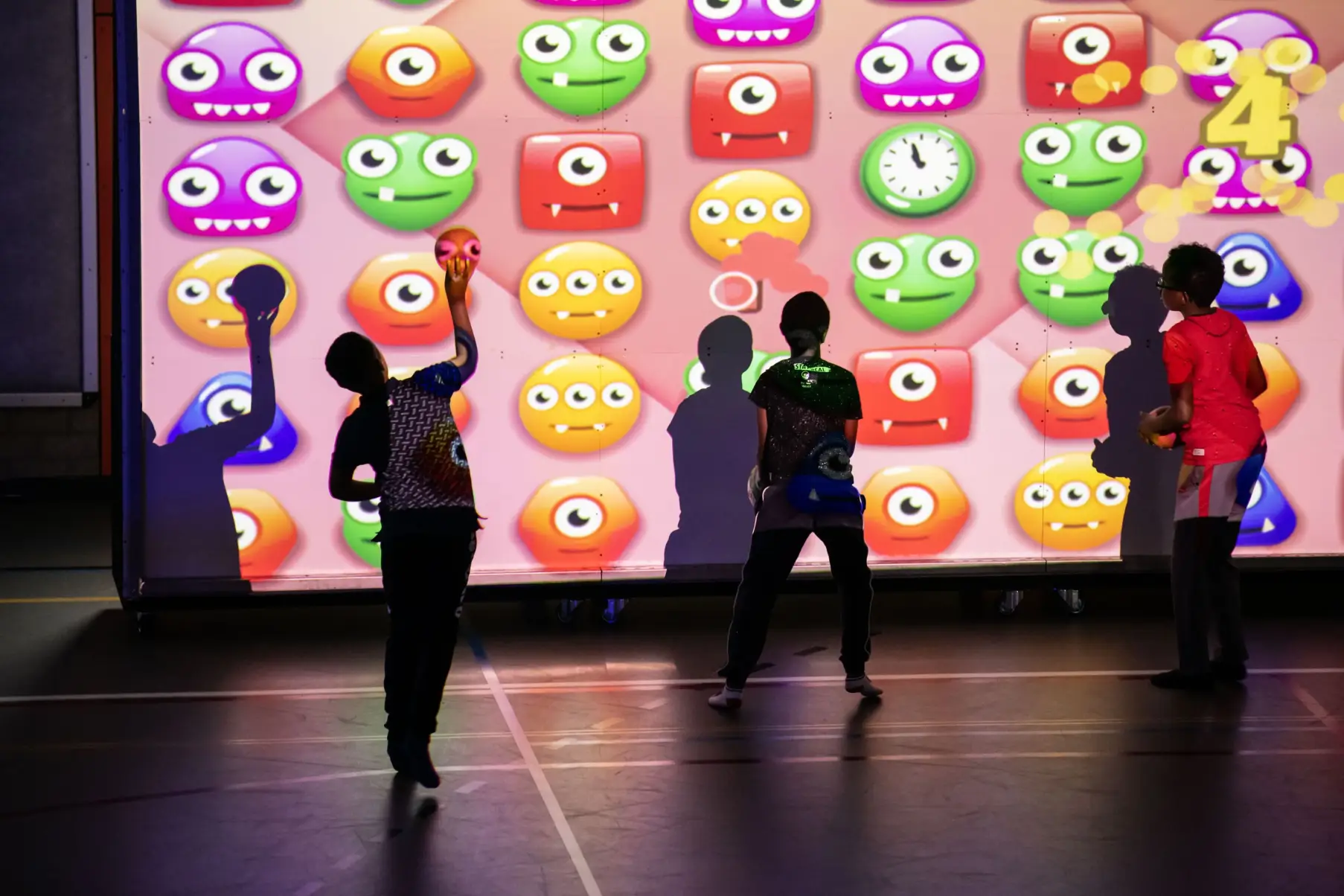 Vier jongeren staan voor een grote interactieve muur met kleurrijke cartoongezichten en gooien ballen tegen het scherm.