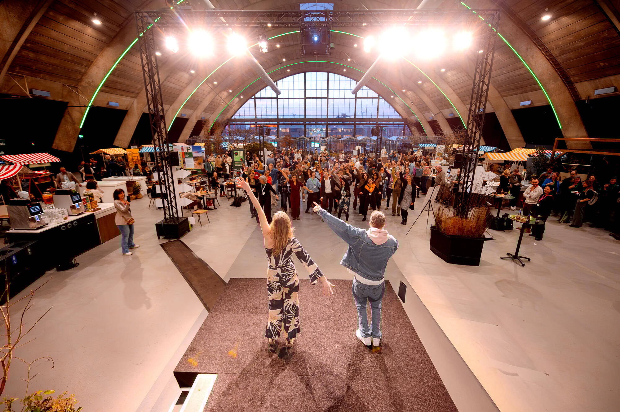 Vrouw en een man staan met de rug naar de camera toe naar een publiek met mensen op de Natuurinclusief Manifestatie in de Brabanthallen. Ze zwaaien een hand in de lucht en het publiek ook.