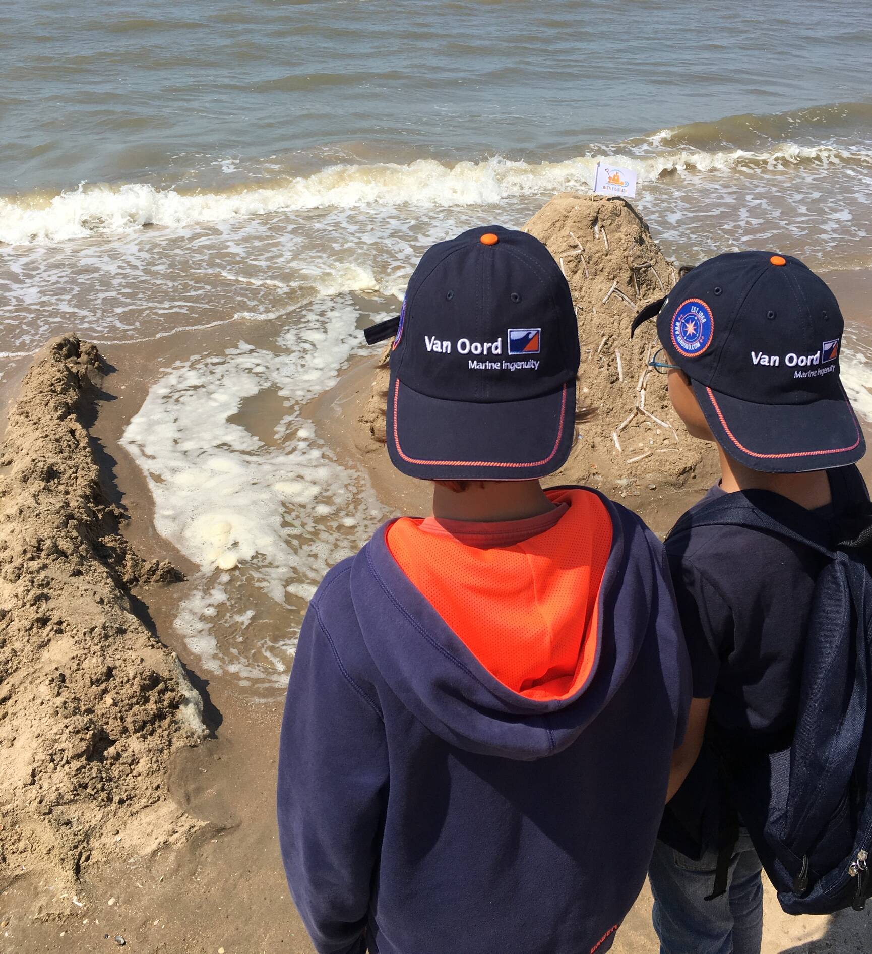 Two kids playing with Sand at the beach, both wearing a Van Oord cap