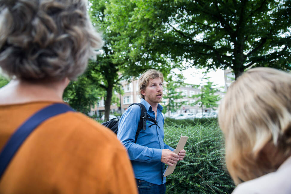 Een vrijwillige gids vertelt een groep mensen wat over natuur in de stad. Ze staan bij een plantsoen.